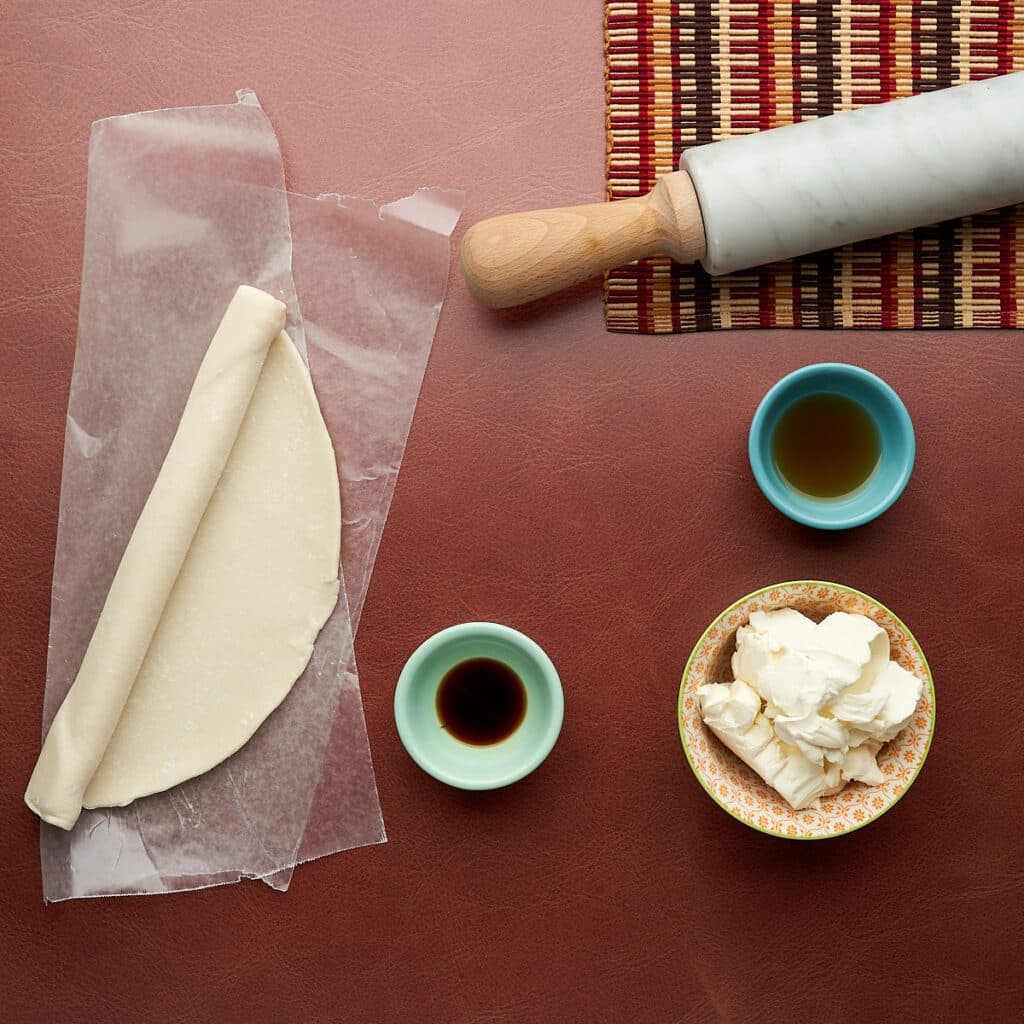 Pre-made pie dough and cream cheese on a leather surface with a rolling pin and placemat on the top right corner